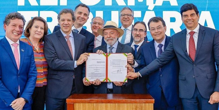 Na fotografia, o Presidente Luis Inácio Lula da Silva, o ministro Fernando Haddad, e outros políticos, seguram um arquivo que sanciona a Reforma Tributária no Brasil.